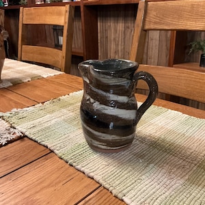 May include: A handmade ceramic pitcher with a dark brown, white, and black swirl design. The pitcher has a handle and a spout. It is sitting on a green and white woven table runner on a wooden table. The background includes wooden shelves and chairs.