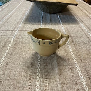 May include: A tan ceramic pitcher with a handle and spout, decorated with blue geometric designs and the number "1822". The pitcher is sitting on a striped tablecloth.