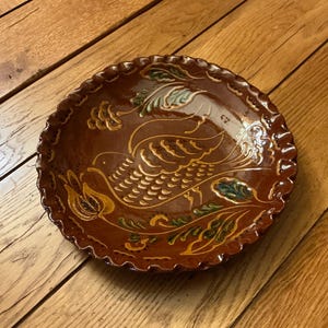 May include: A brown ceramic plate with a ruffled edge. The plate features a hand-painted design of a bird with gold, green, and yellow accents. The bird is surrounded by floral and leaf motifs. The plate is set on a wooden surface.