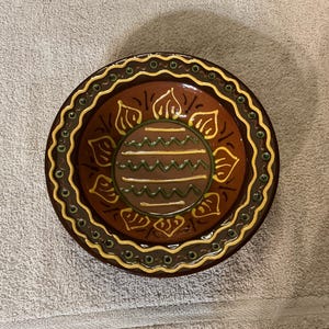 May include: A round, decorative ceramic plate with a brown base and intricate designs. The plate features a central pattern with horizontal lines and leaf-like shapes, accented by yellow and green details. The outer rim has a repeating pattern.