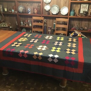May include: A handmade quilt with a dark green background and a repeating pattern of colorful squares. The squares are in shades of pink, yellow, brown, and white. A red border frames the quilt, which is draped over a wooden table.