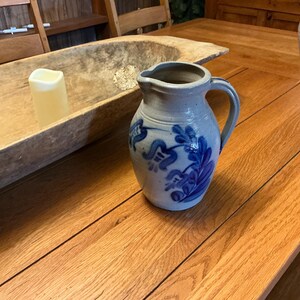 May include: A stoneware pitcher with a handle and a spout, featuring blue floral designs on a light gray background. The pitcher is sitting on a wooden table.