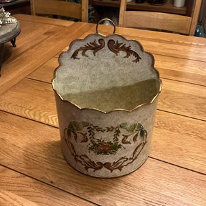 May include: A semi-circular wall-mounted metal pocket with a scalloped top edge and a hanging loop. The off-white surface features a distressed texture, gold-toned trim, and a central floral motif with green leaves and small red flowers.