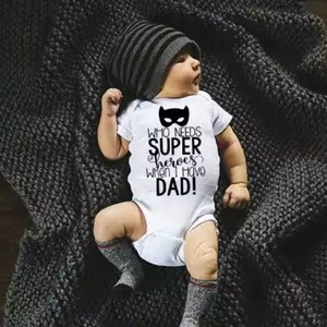 Könnte beinhalten: Ein weißer Baby-Strampler mit schwarzem Text, der "Wer braucht Superhelden, wenn ich Papa habe!" lautet. Der Strampler hat ein schwarzes Masken-Motiv über dem Text. Ein Baby trägt den Strampler und eine schwarz-weiß gestreifte Mütze. Das Baby liegt auf einer grauen Strickdecke.