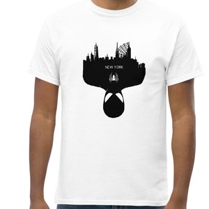 May include: White t-shirt featuring a black silhouette of the New York City skyline, with the text "NEW YORK" and a spider symbol. The design includes a Spider-Man mask. The shirt is made of a soft, comfortable material.