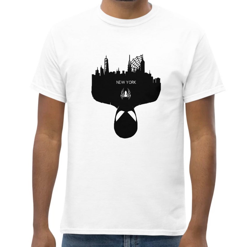 May include: White t-shirt featuring a black silhouette of the New York City skyline, with the text "NEW YORK" and a spider symbol. The design includes a Spider-Man mask. The shirt is made of a soft, comfortable material.