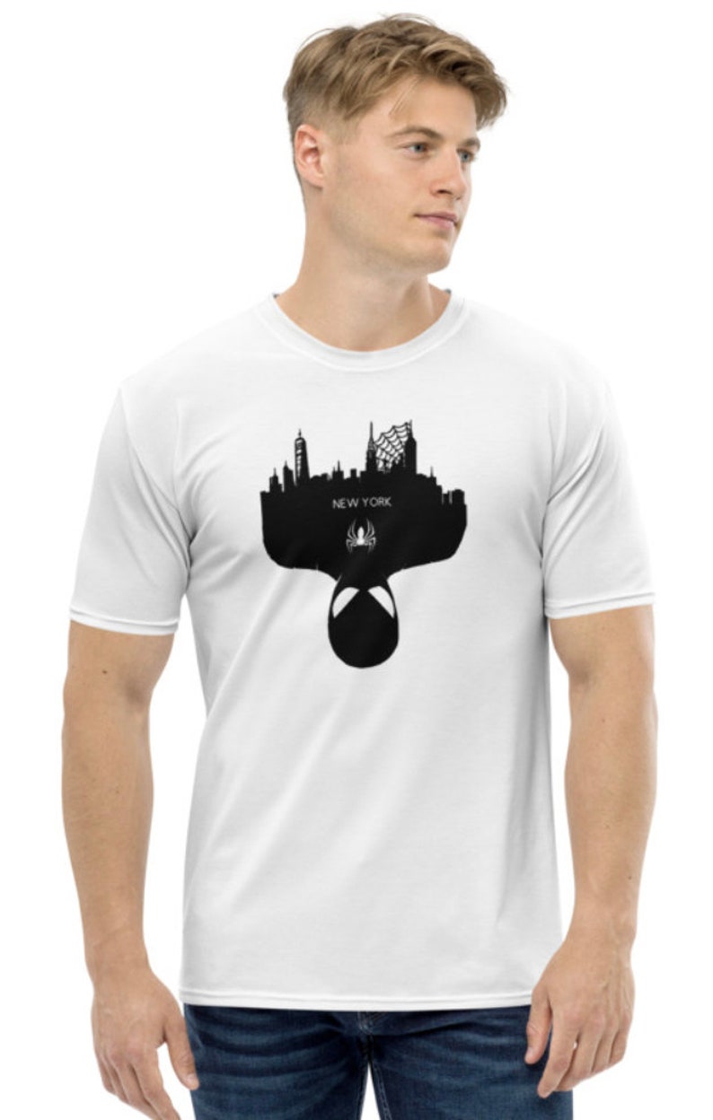 May include: White t-shirt featuring a black silhouette of the New York City skyline, a spider symbol, and the word "NEW YORK". The design includes the mask of a superhero. The shirt is made of a soft, comfortable material.