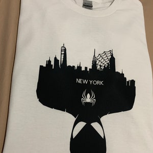 May include: White t-shirt featuring a black silhouette of the New York City skyline, the text "NEW YORK", and a spider symbol. The design includes a spider-man mask silhouette at the bottom.