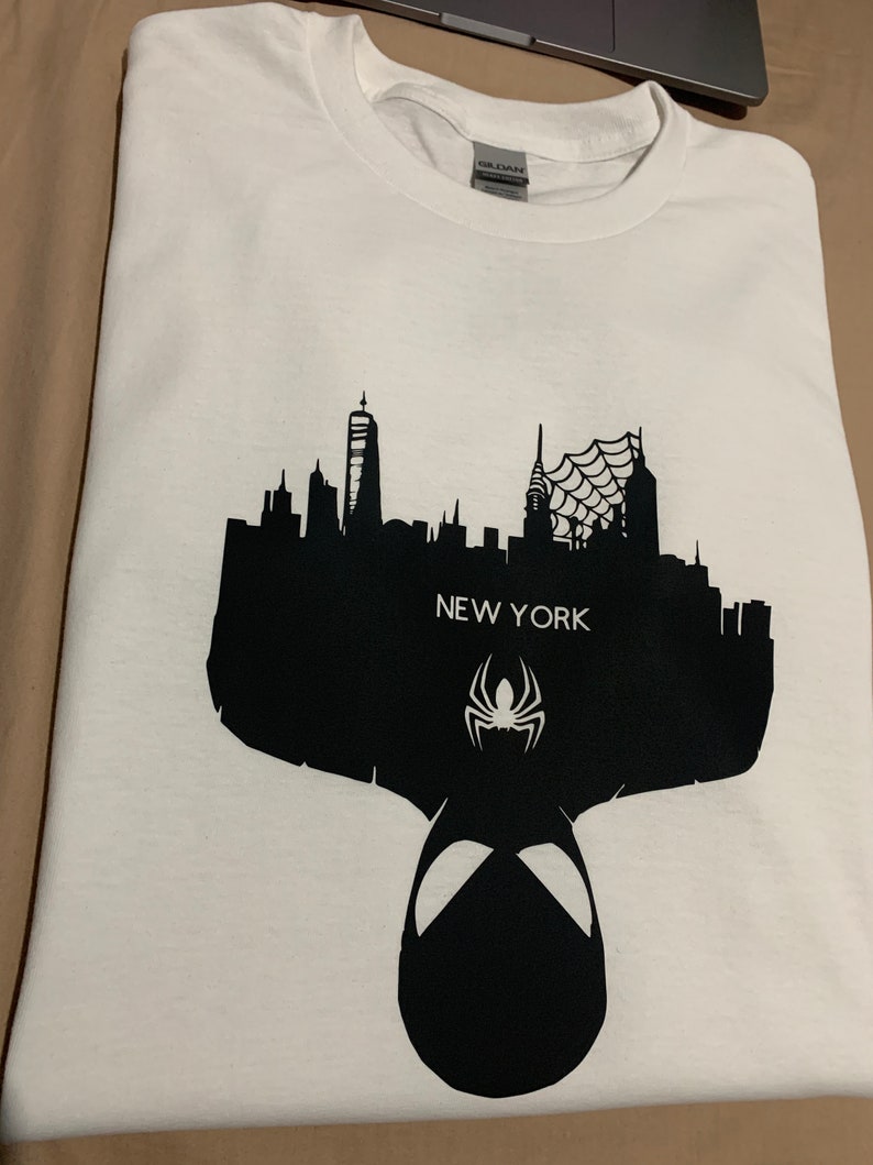 May include: White t-shirt featuring a black silhouette of the New York City skyline, the text "NEW YORK", and a spider symbol. The design includes a spider-man mask silhouette at the bottom.