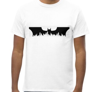 May include: A white t-shirt featuring a black silhouette of the Batman logo, with a bat and a city skyline in the background.
