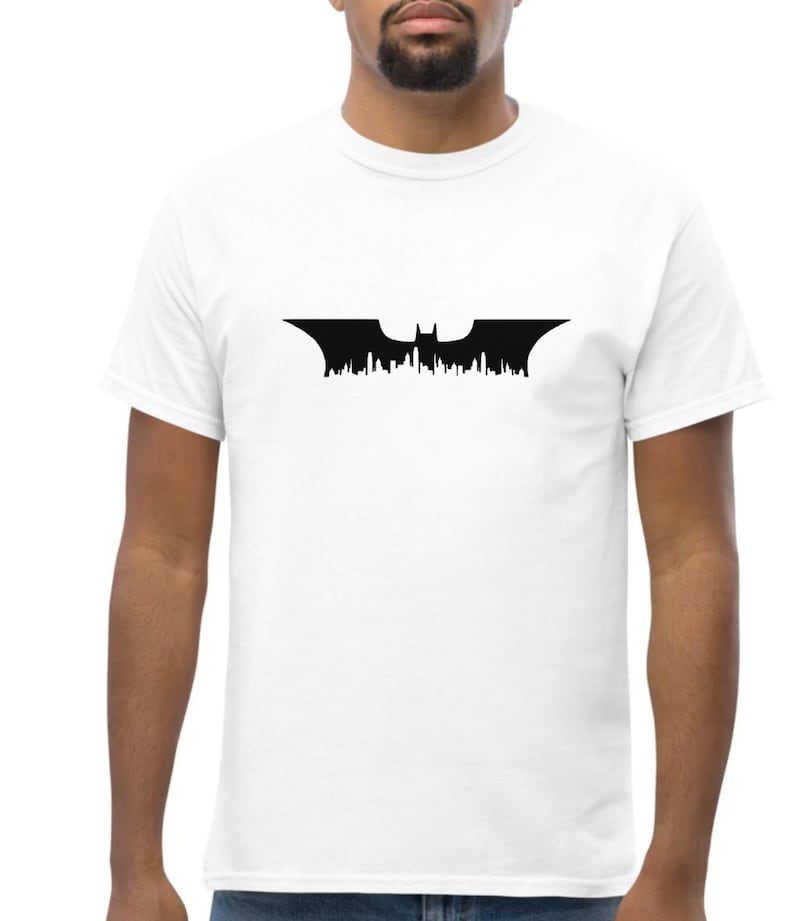 May include: A white t-shirt with a black Batman logo featuring a bat silhouette with a cityscape in the background.