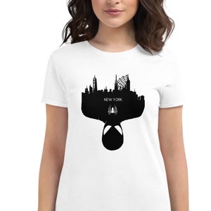 May include: White t-shirt featuring a black silhouette of the New York City skyline, a spider web, and a spider symbol. The word "NEW YORK" is printed in white. The bottom of the design shows the mask of a superhero.