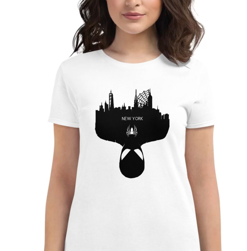 May include: White t-shirt featuring a black silhouette of the New York City skyline, a spider web, and a spider symbol. The word "NEW YORK" is printed in white. The bottom of the design shows the mask of a superhero.