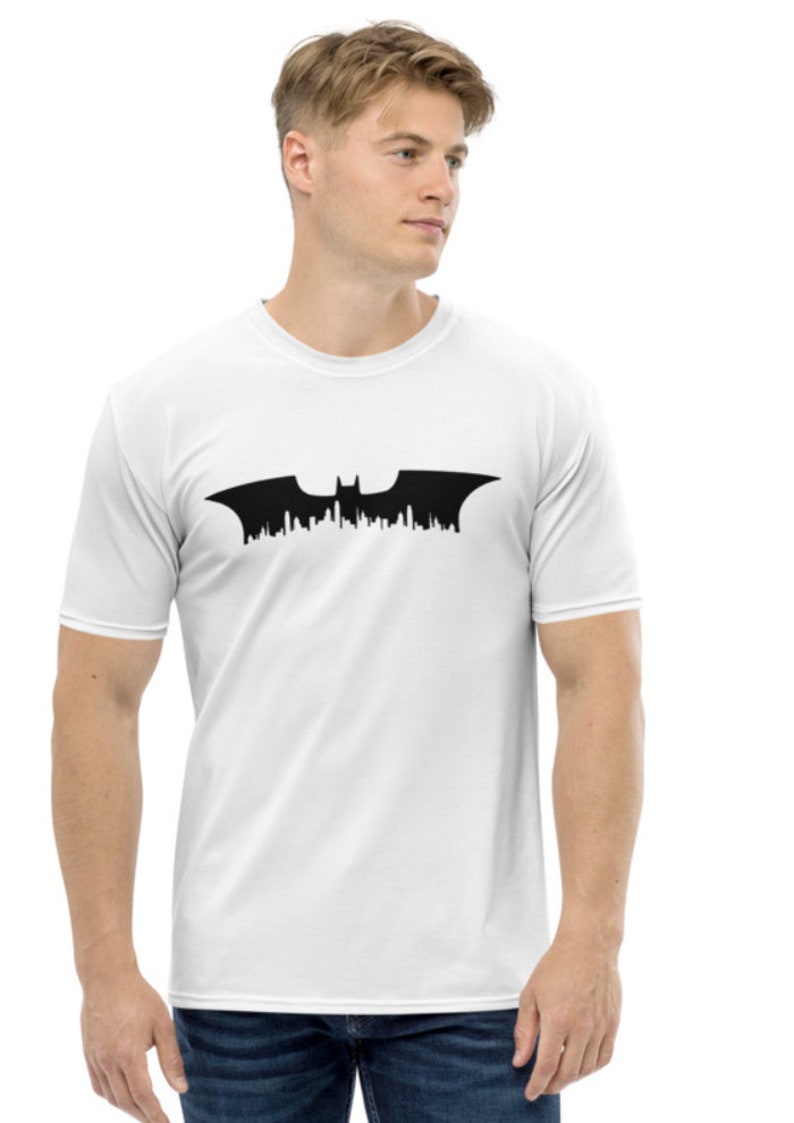 May include: White t-shirt featuring a black silhouette of the Batman logo, with a cityscape design.