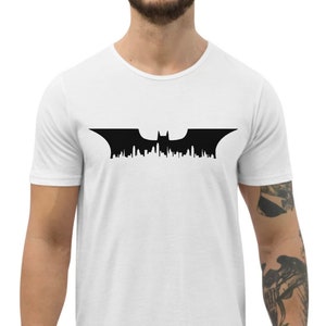 May include: A white t-shirt featuring a black silhouette of the Batman logo, with a cityscape design.