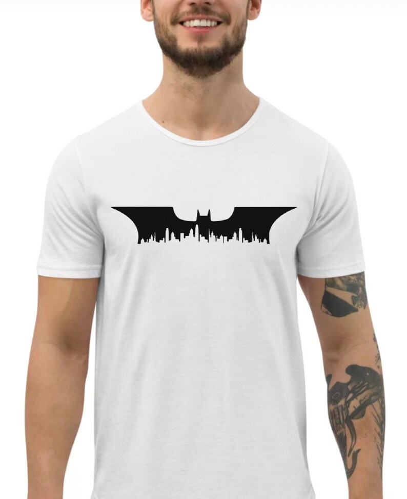 May include: A white t-shirt featuring a black silhouette of the Batman logo, with a cityscape design.