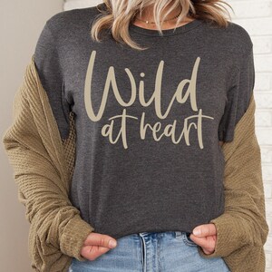 Puede incluir: Una camiseta gris oscuro con el texto "Wild at heart" en color crema.