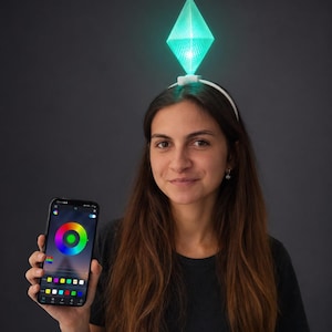 May include: A white headband featuring a translucent, diamond-shaped geometric topper that glows with a bright green light. The device is controlled by a smartphone app displaying a circular color wheel interface for adjusting the light settings.