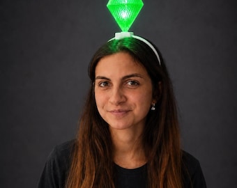 Puede incluir: Un plumbob de color verde brillante, translúcido y con forma de diamante, sujeto a una diadema blanca fina. El objeto geométrico facetado presenta una superficie texturizada y emite una luz brillante, colocado sobre la diadema.