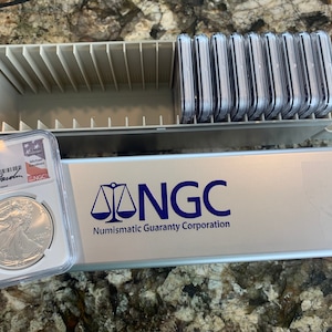 May include: A silver coin in a clear plastic case with a white label that reads "2021 Eagle $1, Eagle Landing T-2, First Day of Issue, MS 70, 5123600-101, Numismatic Guaranty Corporation, NGC". The case is sitting on top of a silver box with the text "NGC, Numismatic Guaranty Corporation" in blue letters. The box has a scale symbol in blue.