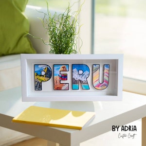 May include: White framed shadow box with the word "PERU" spelled out in large letters. Each letter is filled with a different image of a Peruvian landscape. The box is sitting on a white table with a yellow notebook in front of it.