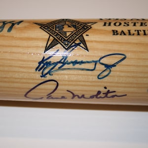 Pode incluir: Um taco de beisebol de madeira com assinaturas de vários jogadores de beisebol, incluindo "Cal Ripken Jr." e "Dave Winfield". O taco também apresenta um logotipo em forma de estrela com o texto "1993 All-Star Game Hosted by Baltimore, July 10-11".