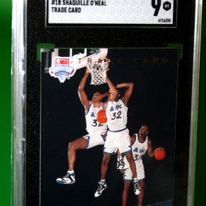 May include: A 1992-93 Upper Deck Shaquille O'Neal basketball trade card, graded 9 MT by SGC. The card features a triple-exposure image of the player in a white and blue pinstriped Orlando Magic uniform, dunking a basketball. The card is encased in a black plastic slab.