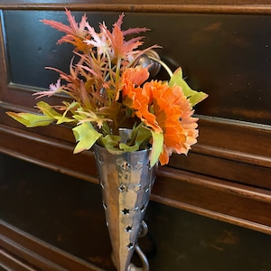 May include: A silver metal vase with a star-shaped cut-out design holds a bouquet of orange flowers and green leaves. The vase is attached to a wooden surface.