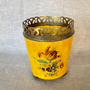 May include: A yellow ceramic planter with a floral design and a gold metal rim. The planter has three small feet and is decorated with a floral pattern in shades of red, blue, and yellow.