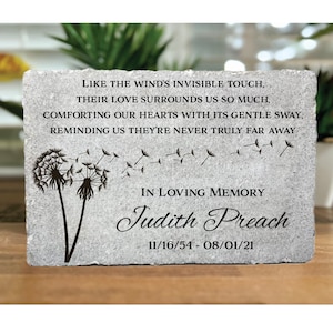 May include: A gray stone memorial with a dandelion design and the text "LIKE THE WIND'S INVISIBLE TOUCH, THEIR LOVE SURROUNDS US SO MUCH, COMFORTING OUR HEARTS WITH ITS GENTLE SWAY, REMINDING US THEY'RE NEVER TRULY FAR AWAY IN LOVING MEMORY Judith Preach 11/16/54 - 08/01/21"
