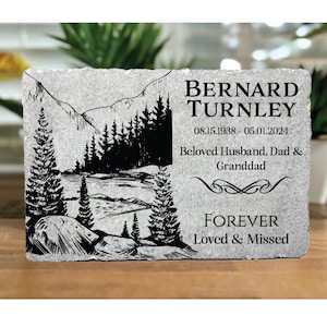 May include: A gray stone memorial with a black and white illustration of a mountain landscape. The text reads "Bernard Turnley 08.15.1938 - 05.01.2024 Beloved Husband, Dad & Granddad Forever Loved & Missed"