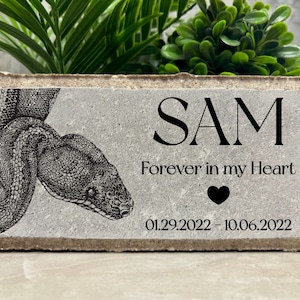 May include: A gray concrete memorial block with a black and white illustration of a snake. The block has the name "SAM" engraved on it, along with the text "Forever in my Heart" and the dates "01.29.2022 - 10.06.2022".