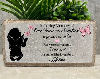 Può includere: Una pietra commemorativa in cemento grigio con una silhouette nera di un bambino angelo che tiene una farfalla. La pietra ha il testo "In Loving Memory of Our Precious Angelica September 15th 2022 You were carried for a Moment but you will be loved for a Lifetime".