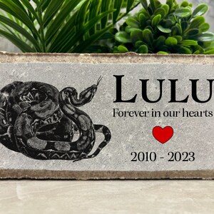 May include: A gray concrete memorial stone with a black and white image of a snake, the name "LULU" in black text, the phrase "Forever in our hearts" in black text, a red heart, and the dates "2010 - 2023" in black text.
