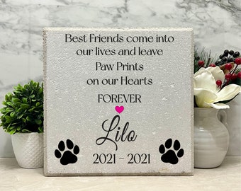 May include: A white stone memorial with the text "Best Friends come into our lives and leave Paw Prints on our Hearts FOREVER" and a pink heart. The name "Lilo" is written in cursive below the heart. The dates "2021 - 2021" are below the name. Two black paw prints are on either side of the dates.