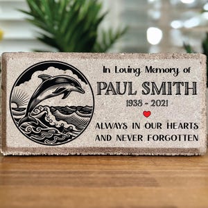 May include: A rectangular stone memorial with a dolphin illustration and the text "In Loving Memory of Paul Smith 1938-2021 Always in Our Hearts and Never Forgotten."