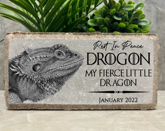May include: A gray stone memorial with a black and white illustration of a bearded dragon. The text reads "Rest in Peace Drogon My Fierce Little Dragon January 2022".
