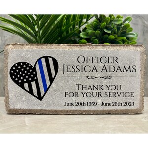 May include: A gray concrete memorial block with a blue and white heart design. The text reads "OFFICER JESSICA ADAMS THANK YOU FOR YOUR SERVICE June 20th 1959 - June 26th 2023".