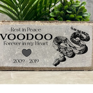 May include: A gray concrete memorial block with a black snake design and the text "Rest in Peace VOODOO Forever in my Heart 2009-2019". A black heart is below the text.