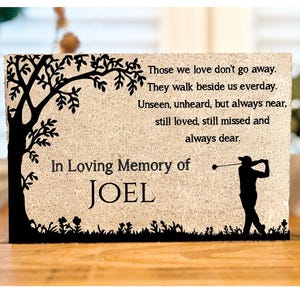 May include: A memorial plaque with a textured beige surface. The black silhouette of a tree and a golfer are on the left and right. Text reads "Those we love don't go away...In Loving Memory of JOEL."