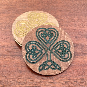 May include: Two round wooden coasters with a Celtic knot design. One coaster features a gold shamrock design, while the other has a green shamrock design.