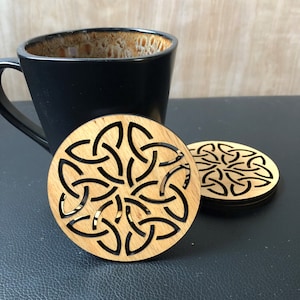 May include: Two round wooden coasters with a Celtic knot design. The coasters are made of light brown wood with a dark brown knot pattern. The coasters are on a black surface.