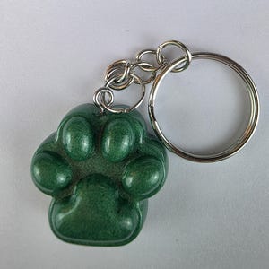 May include: A green paw print keychain with a silver key ring and chain. The paw print is a solid, glossy green color. The keychain is designed to be a pet-themed accessory.