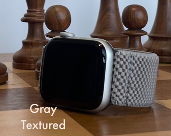 May include: A grey textured Apple Watch band with a silver Apple Watch on a chessboard.