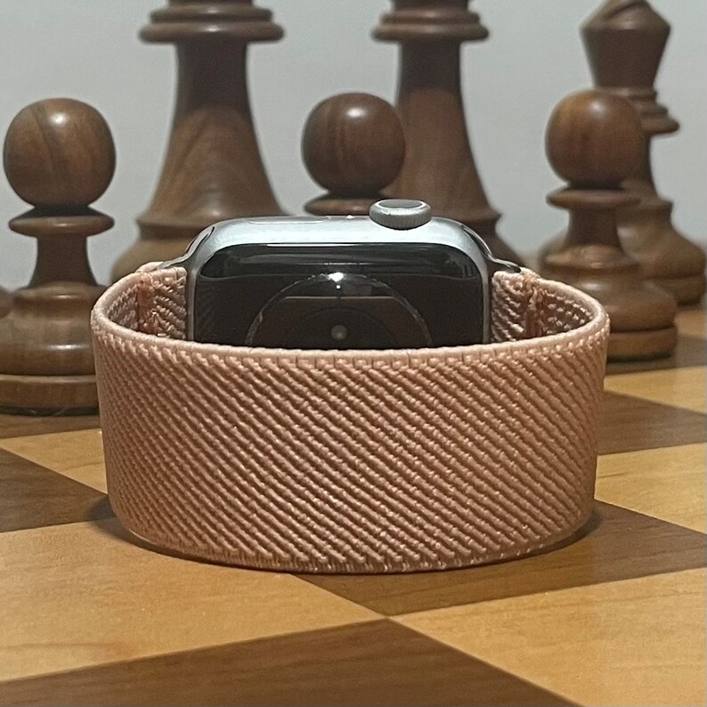 May include: A silver Apple Watch with a light pink woven band. The watch is sitting on a wooden chessboard.