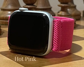 May include: A silver smartwatch with a black screen and a hot pink elastic strap. The text "Hot Pink" is displayed below the watch. The watch is on a wooden surface, with chess pieces in the background.