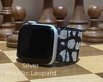 May include: A silver Apple Watch with a black and silver patterned band. The band has a metallic leopard print design. The watch is on a wooden surface.