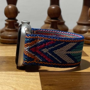 May include: A silver Apple Watch with a colorful woven band. The band has a geometric pattern in shades of blue, orange, pink, and white.