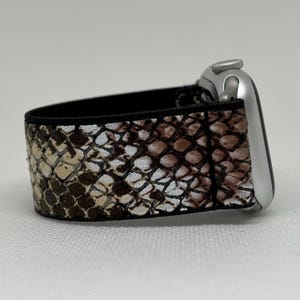 May include: An Apple Watch strap featuring a snakeskin design in brown, beige, and white. The strap is connected to a silver Apple Watch. The strap has a black elastic backing.