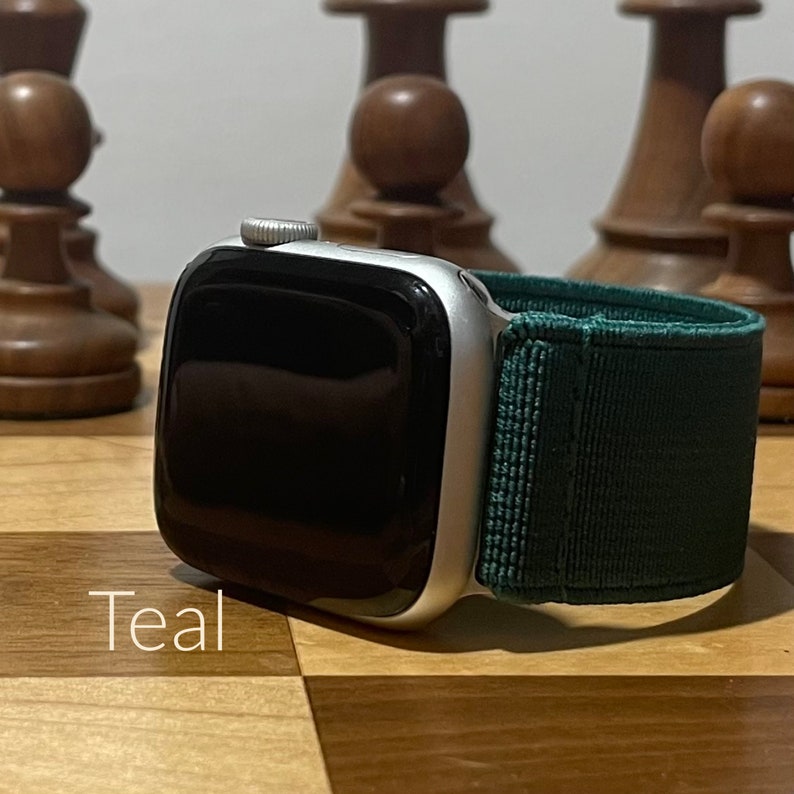 May include: A silver Apple Watch with a teal green fabric band. The watch is on a wooden surface with chess pieces in the background.